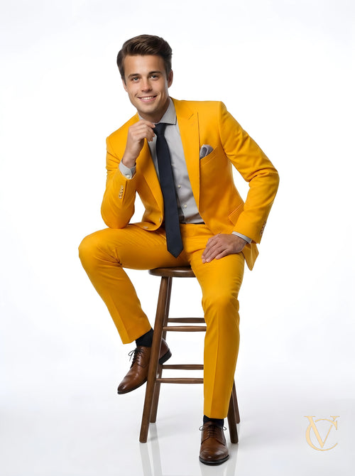 Young man wearing a vibrant 2-Piece Suit - Yellow | Vellure Couture with a tailored fit, paired with a gray shirt, black tie, and brown dress shoes, seated on a wooden stool.
