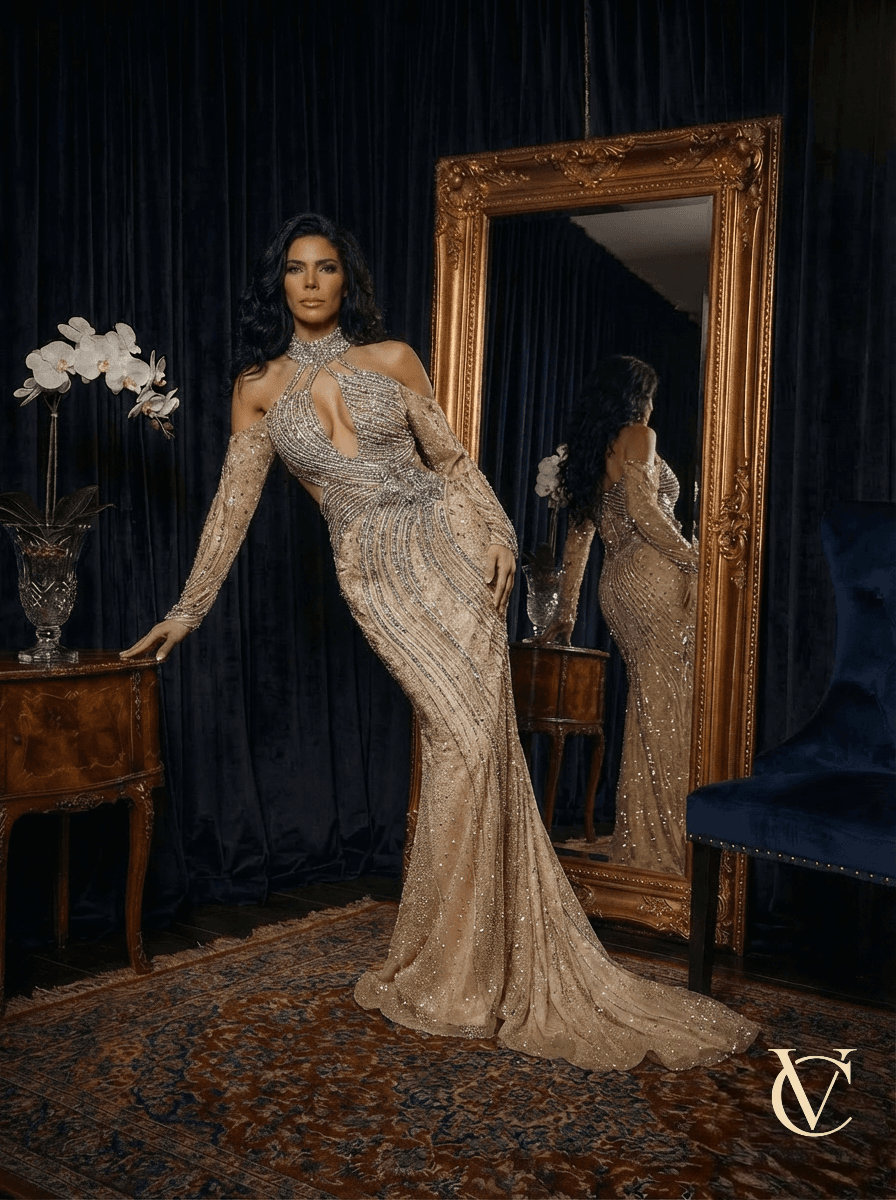 Elegant woman wearing the Satin & Crystals Gown - Champagne | Vellure Couture, a crystal embellished satin evening gown with cut-out detail, standing by a vintage wooden table and large ornate mirror.