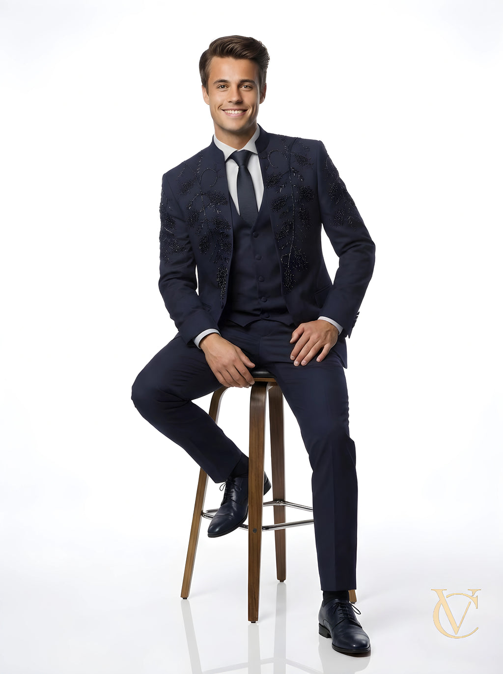 A smiling man wearing a navy tailored 3-Piece Suit | Vellure Couture with subtle black embellishments, seated on a high wooden stool against a white background.