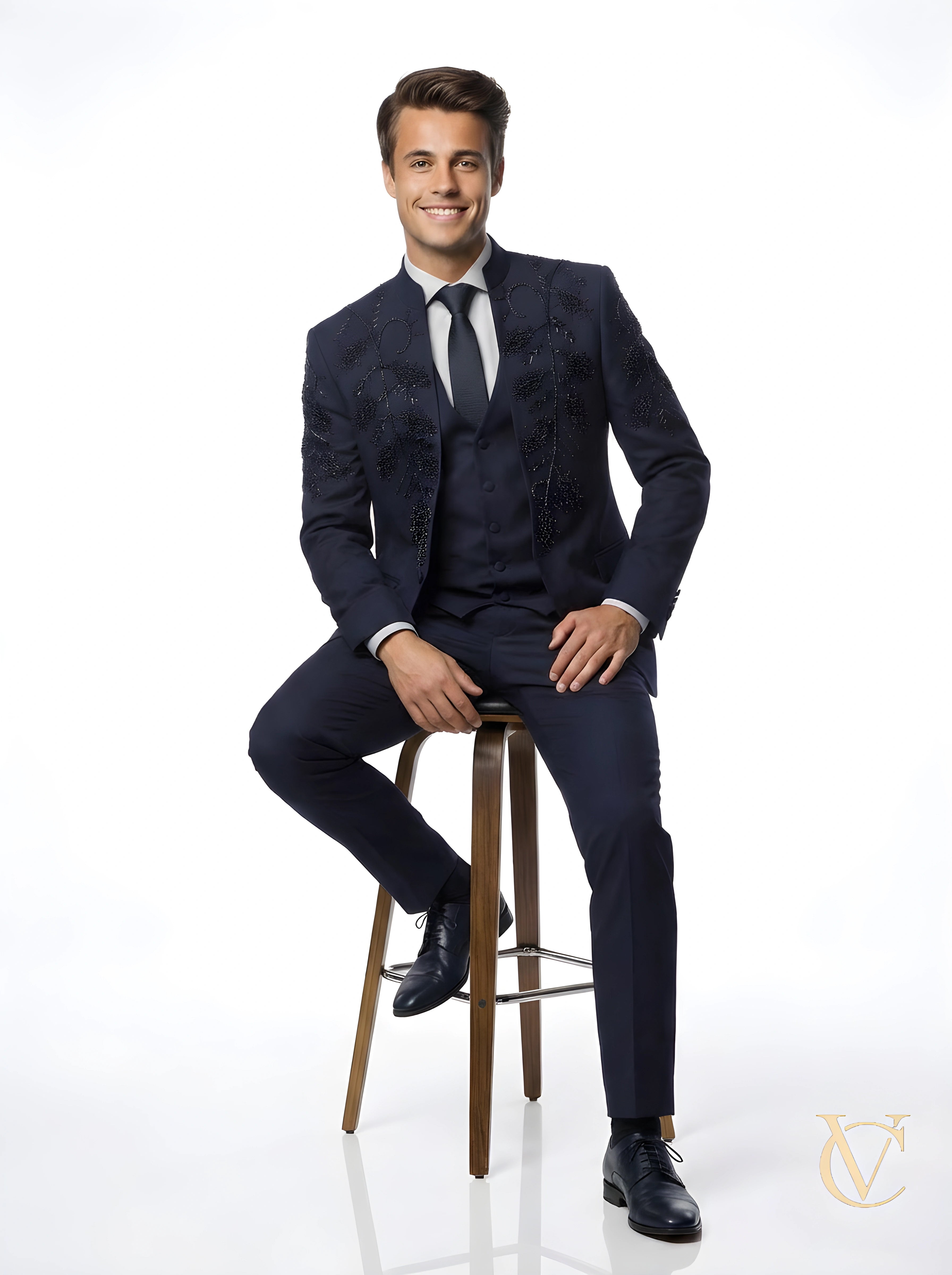 A smiling man wearing a navy tailored 3-Piece Suit | Vellure Couture with subtle black embellishments, seated on a high wooden stool against a white background.