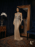Elegant woman wearing Satin & Crystals Gown - Champagne | Vellure Couture, featuring intricate crystal embellishments, a keyhole neckline, and long sheer sleeves, standing in a luxurious room with ornate decor.
