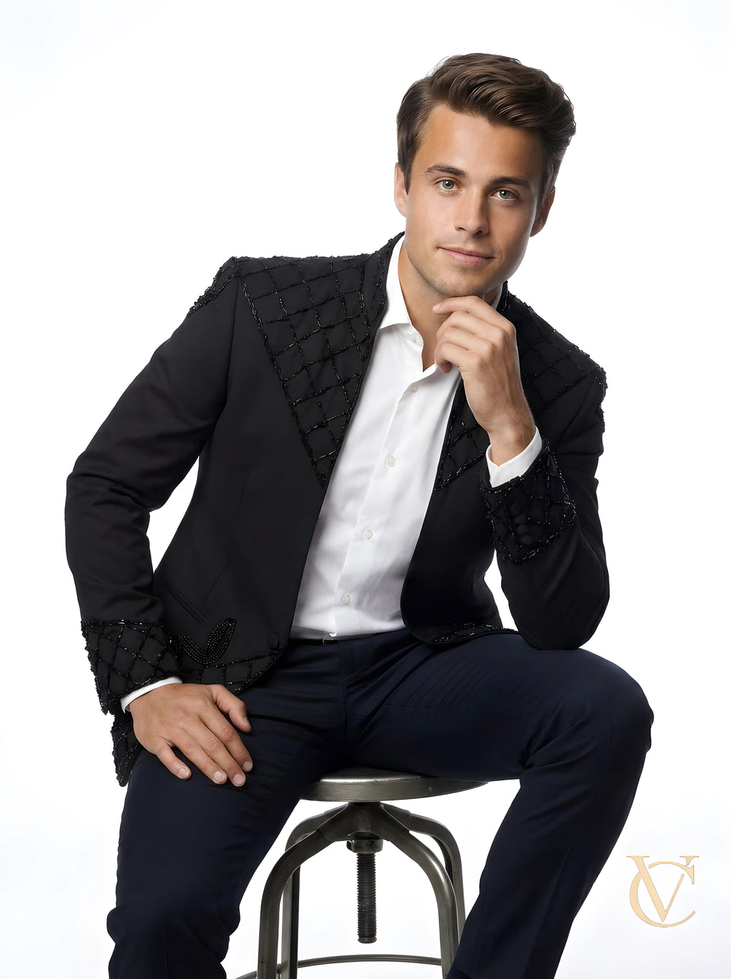 Man wearing a sculpted fit 2-Piece Suit - Black | Vellure Couture with detailed textured lapels and cuffs, paired with a crisp white shirt, seated on a stool against a white background.