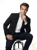 Man wearing a sculpted fit 2-Piece Suit - Black | Vellure Couture with detailed textured lapels and cuffs, paired with a crisp white shirt, seated on a stool against a white background.